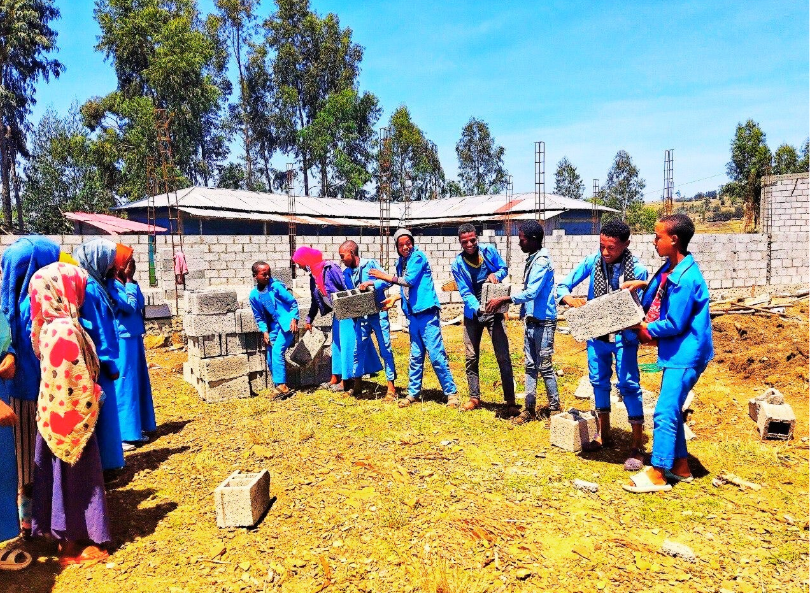 People in blue uniforms lifting bricks at a construction site outdoors on a sunny day.