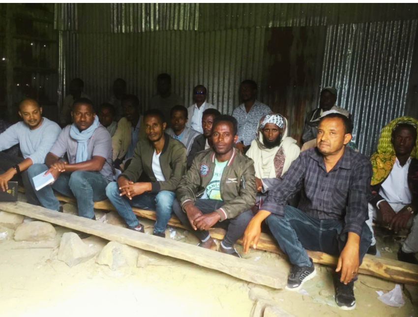 Group of men seated on a wooden bench inside a building with a corrugated metal roof.