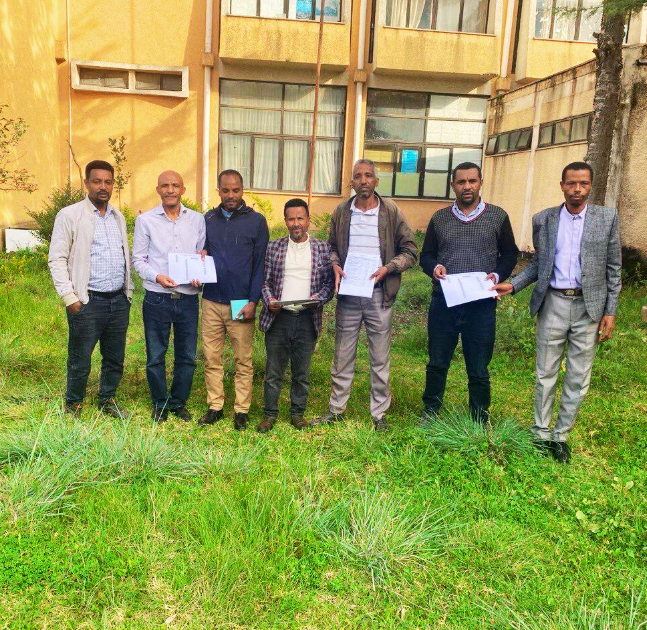 Seven men standing in front of a building, holding papers, on grass.