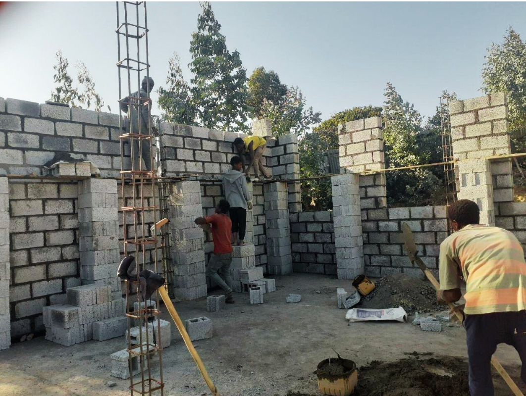 Construction workers building a brick structure outdoors. Men are laying blocks, with tools and materials nearby.