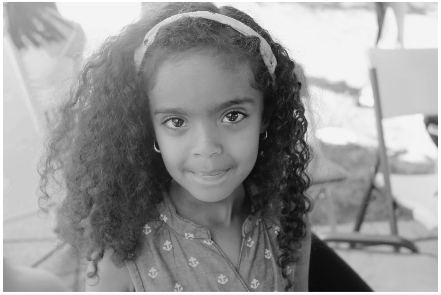Young child with curly hair and a headband smiles, outdoors.