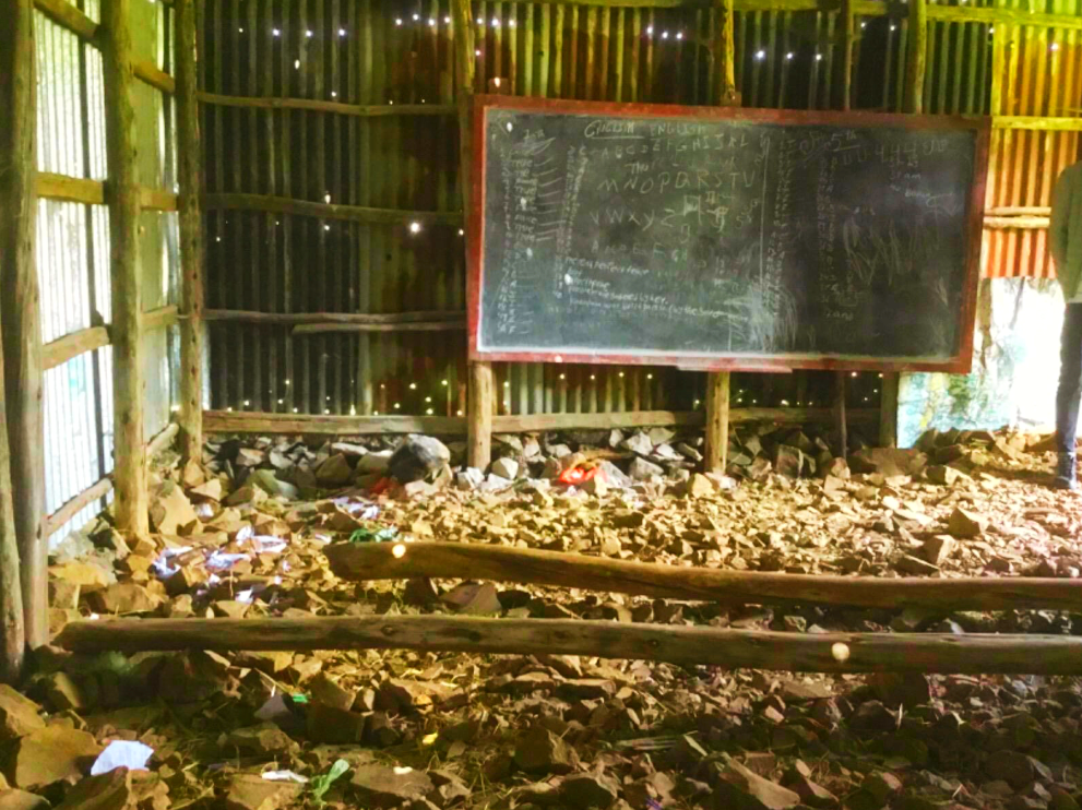 Dilapidated classroom interior with blackboard, rubble, and corrugated metal walls.