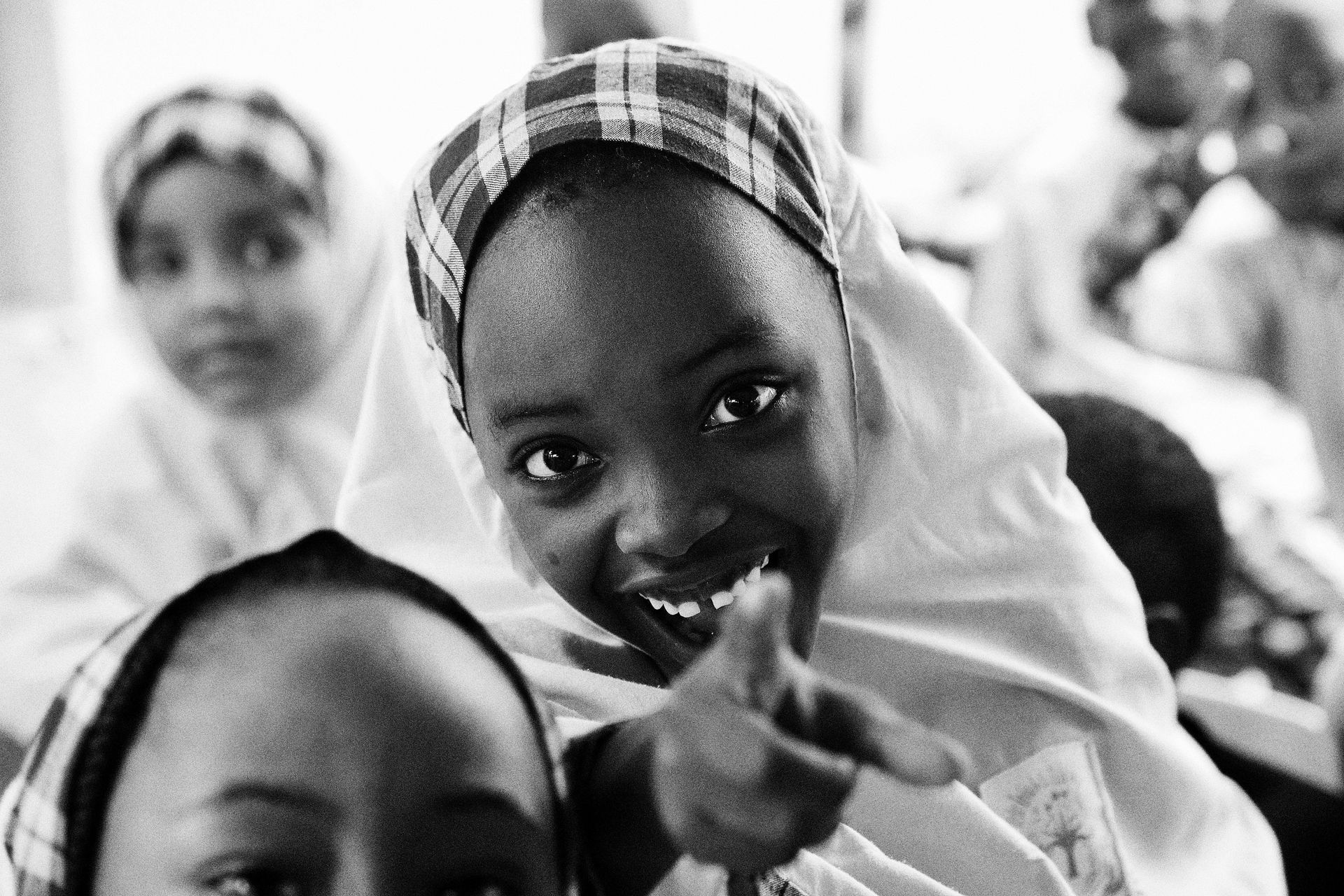 Smiling girl in headscarf points at the camera with other girls in the background.