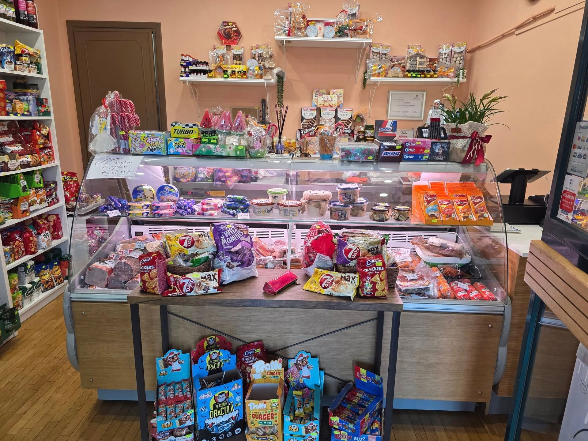 Interno di un negozio con una vetrina piena di snack e dolcetti, scaffali di prodotti e decorazioni.