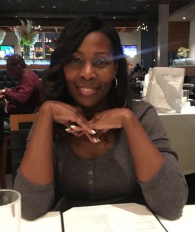 Woman with dark hair smiles, resting chin on crossed hands at a restaurant table.