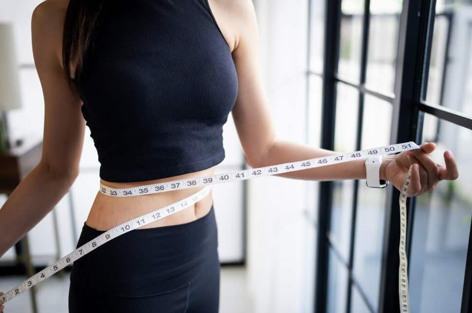 Woman in black athleisure measuring waist with a tape measure near a window.