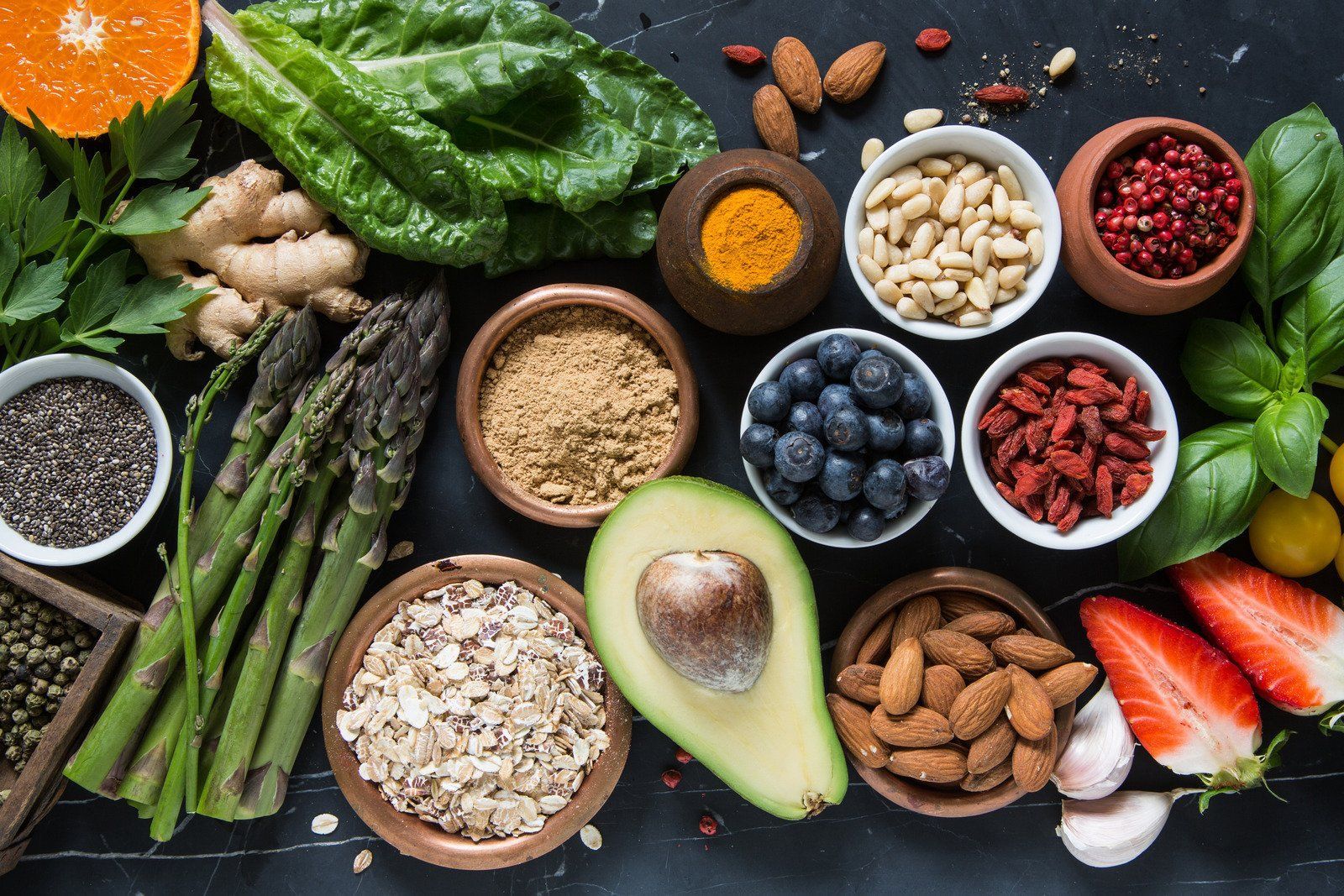Assortment of colorful healthy foods, including fruits, vegetables, nuts, and spices, on a dark surface.