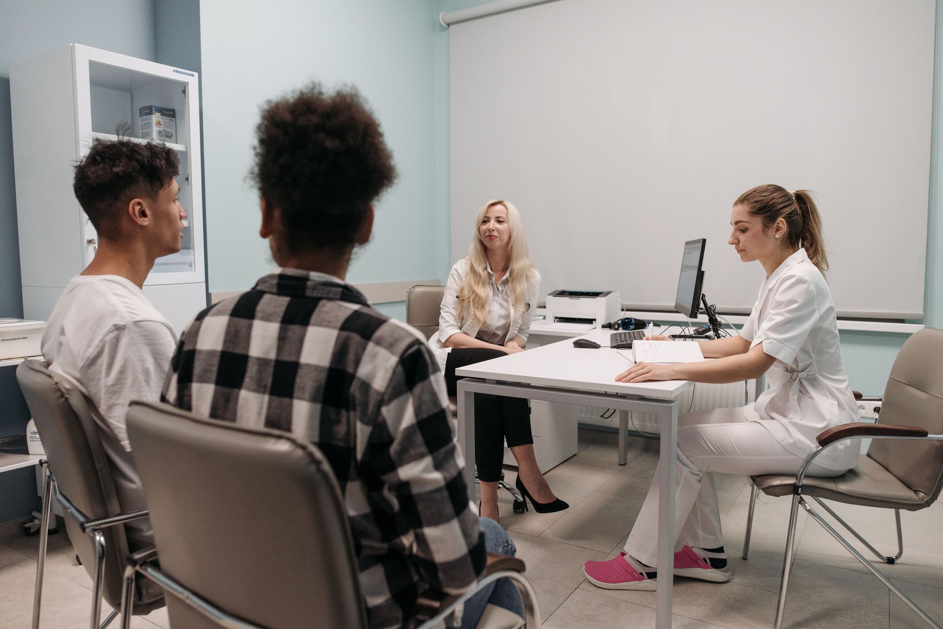 Meeting in a bright office, three people seated around a table with a woman speaking at the front.