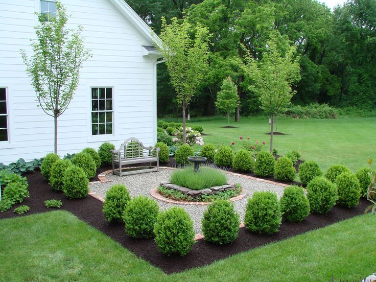 A gravel courtyard with a wooden bench, central birdbath, and symmetrical green shrubs next to a white farmhouse exterior.