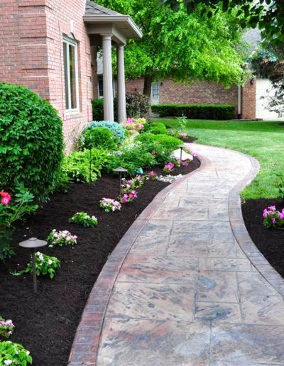 A curving stone walkway lined with small flowering plants, dark mulch, and outdoor lighting leading to a brick house.