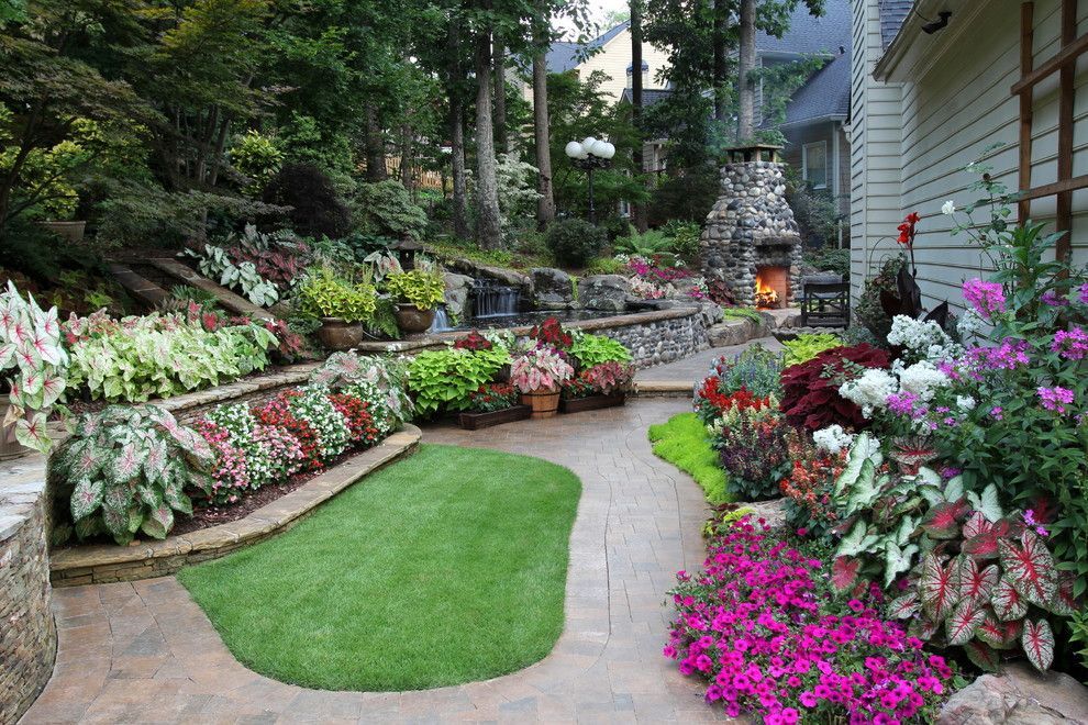 A landscaped patio with a stone fireplace, a lush flowerbed, an artificial grass patch, and a house exterior.