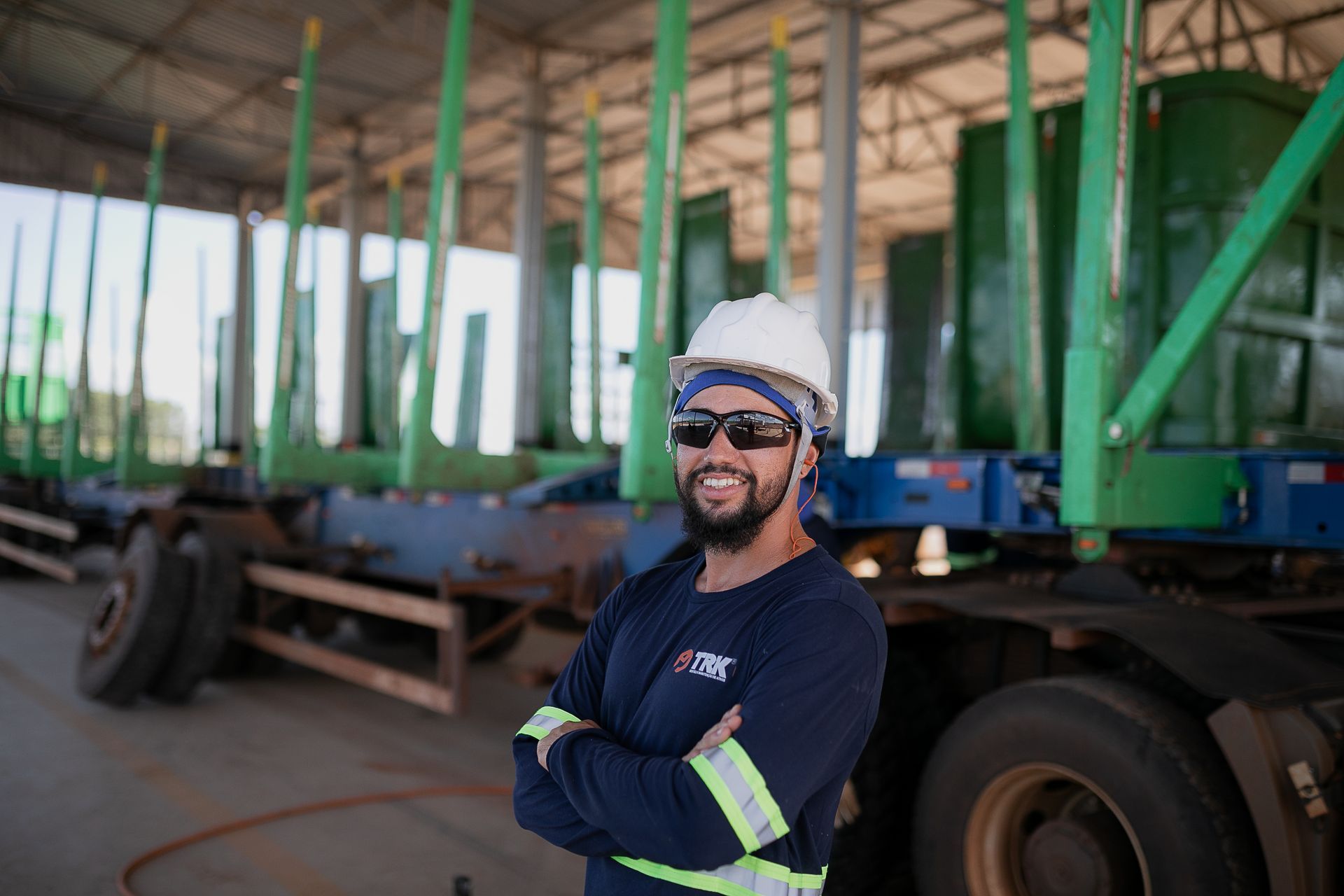 Operário da construção civil, usando capacete e óculos de sol, em pé em frente a uma carreta com equipamentos verdes.