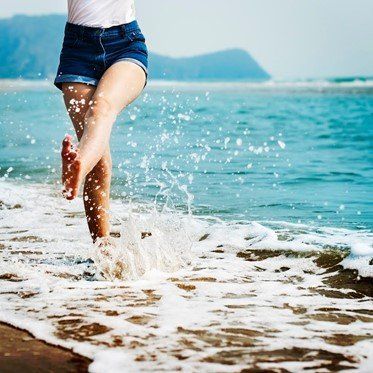 Person running barefoot in the ocean, kicking up water. Wearing denim shorts, a white top, at the beach.