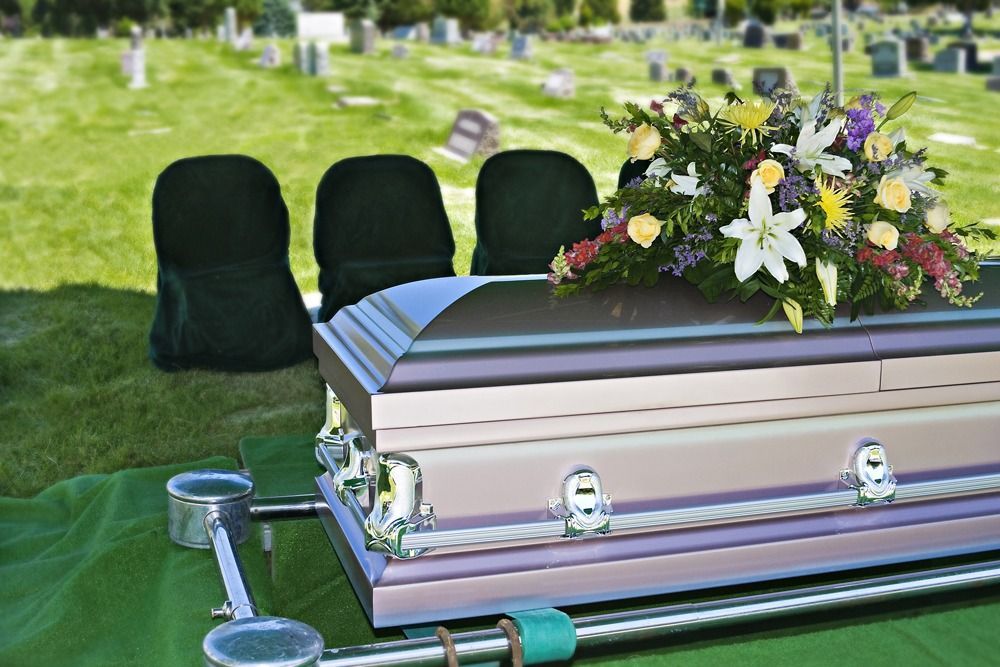 White casket with floral spray at a graveside, with chairs and green grass in the background
