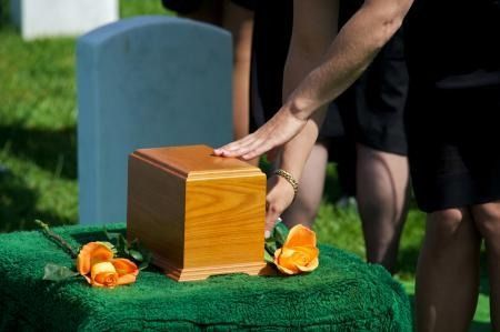 Hand resting on a polished wooden urn beside orange flowers at a gravesite