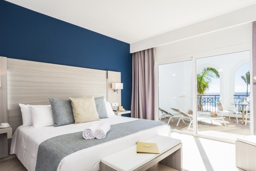 Camera d'albergo con letto king-size, parete blu, balcone con vista sull'oceano e palme.