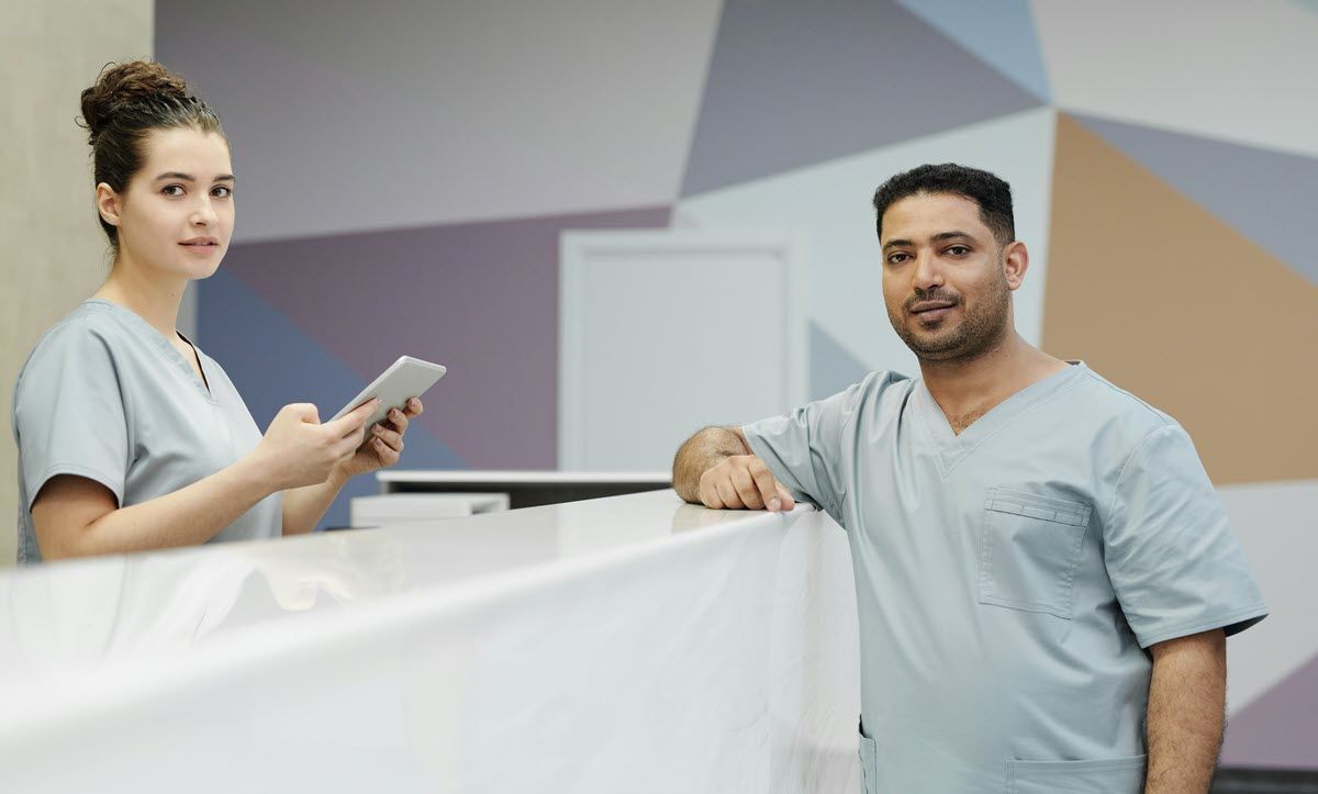 Two healthcare professionals in light blue scrubs standing behind a medical reception desk in a modern office.