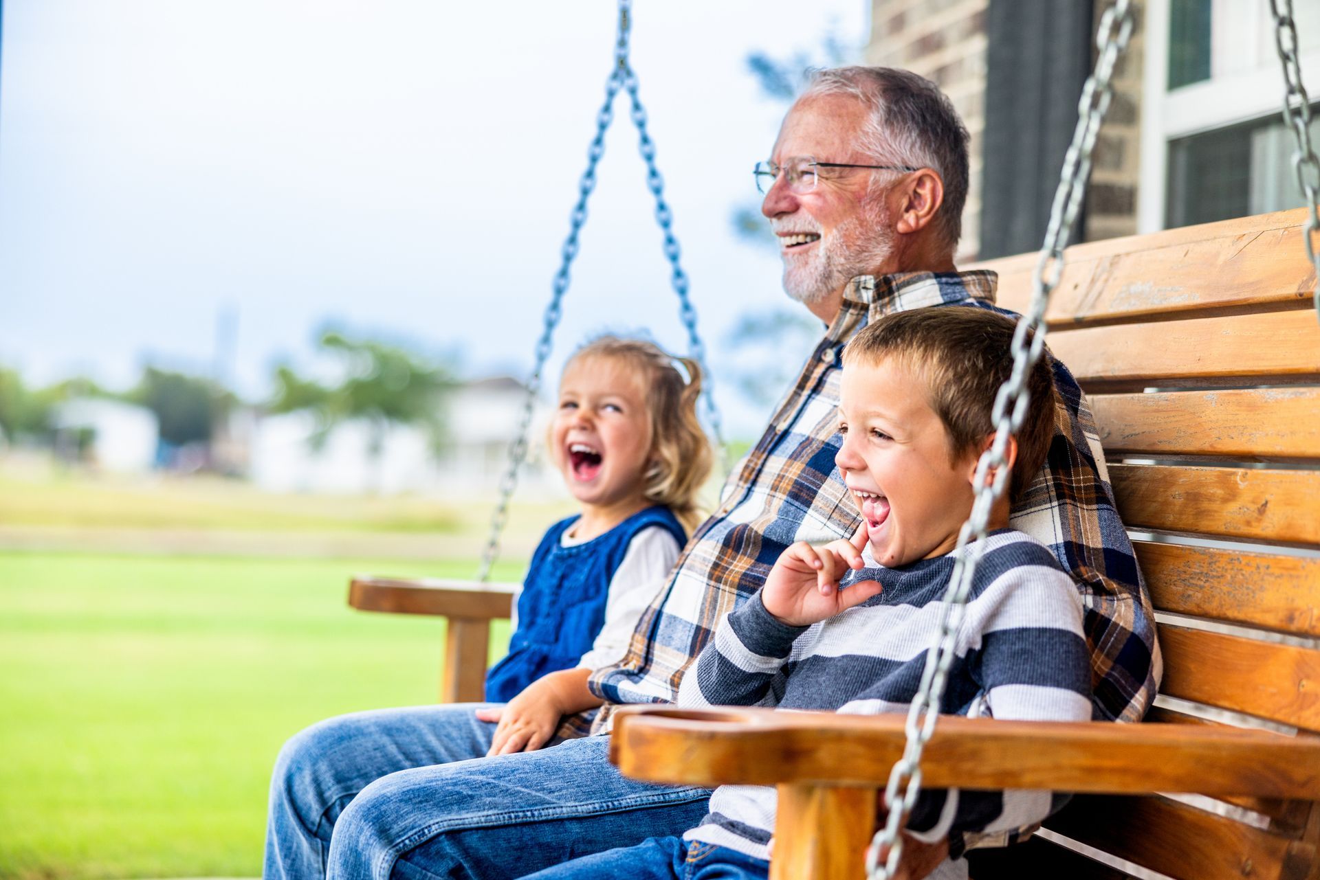 An older adult and two children laugh while sitting together on a wooden porch swing outdoors.