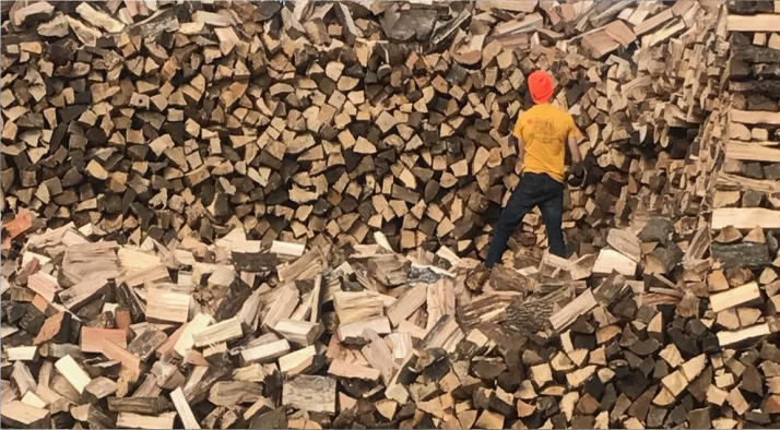 Person in red hat and yellow jacket climbing over a large pile of chopped firewood.