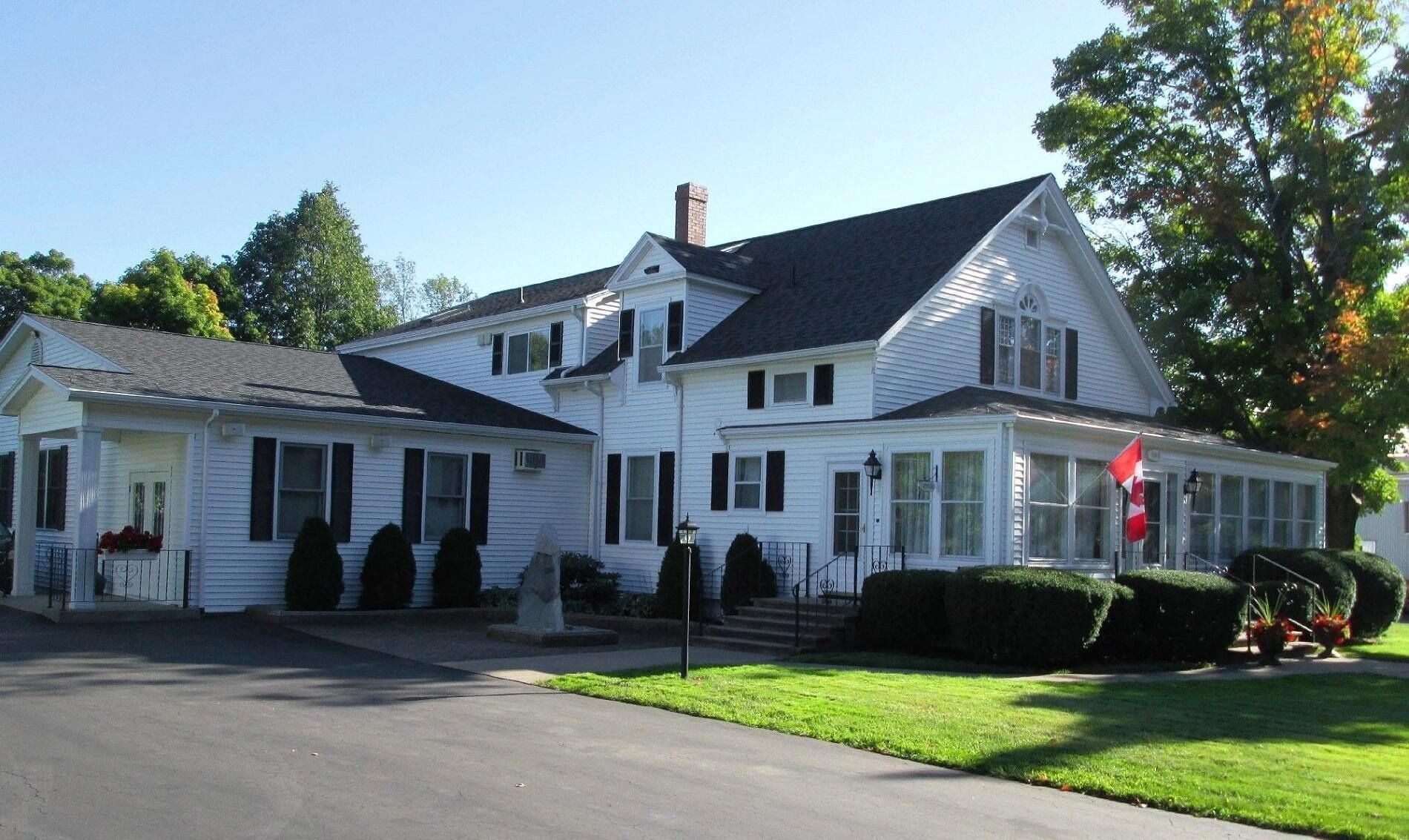 A large, two-story white house with dark shutters, a front porch, and a driveway, surrounded by green lawns and trees.