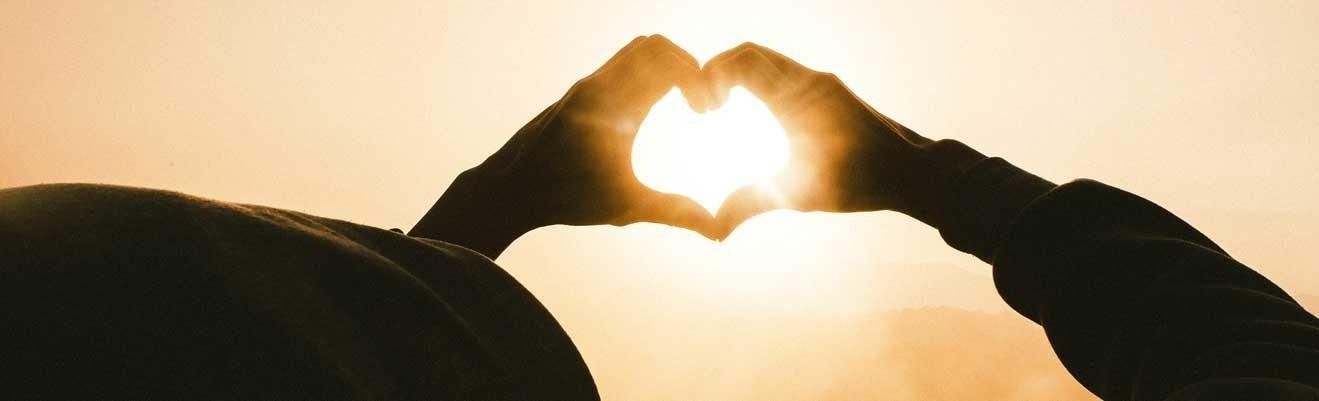 Hands shaped into a heart silhouette against a bright, warm golden sunset.
