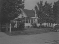 A black and white photograph of a two-story suburban house with a wrap-around porch, surrounded by trees and a car nearby.