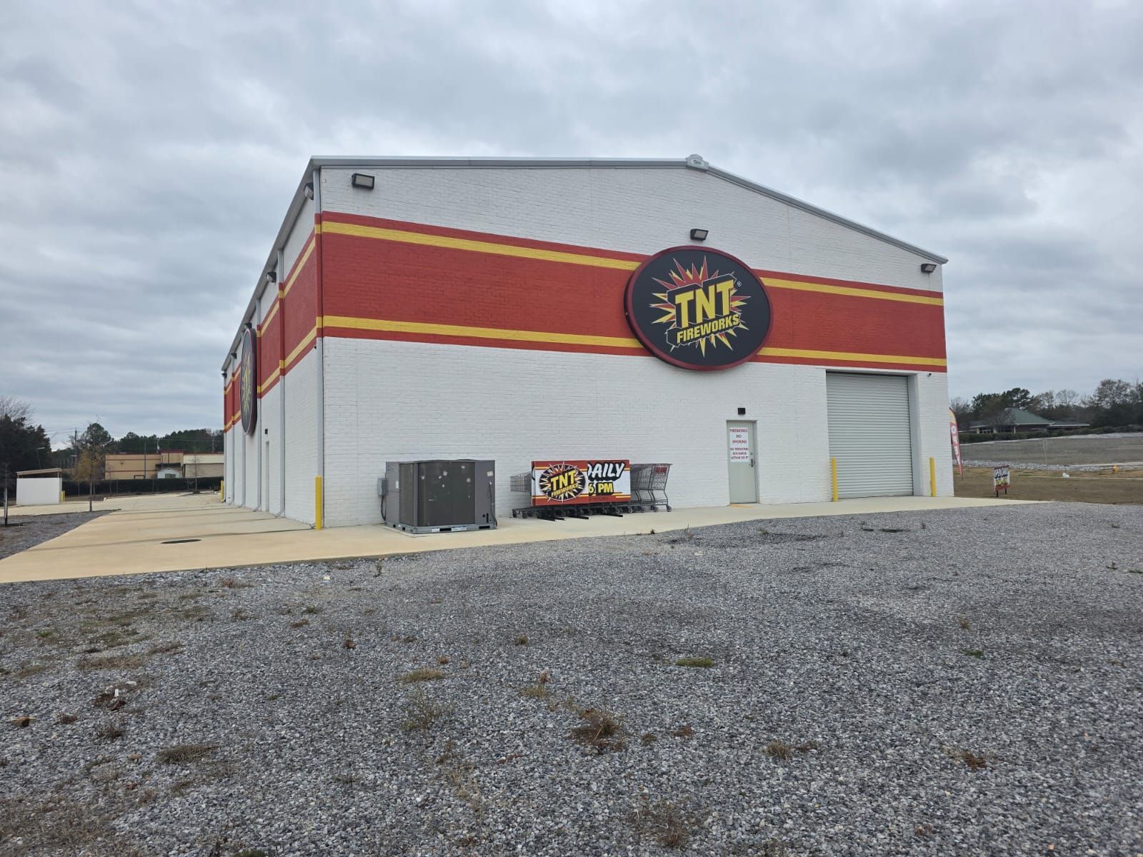 Large white building with red and yellow stripes and a round sign, viewed from a gravel lot under cloudy sky