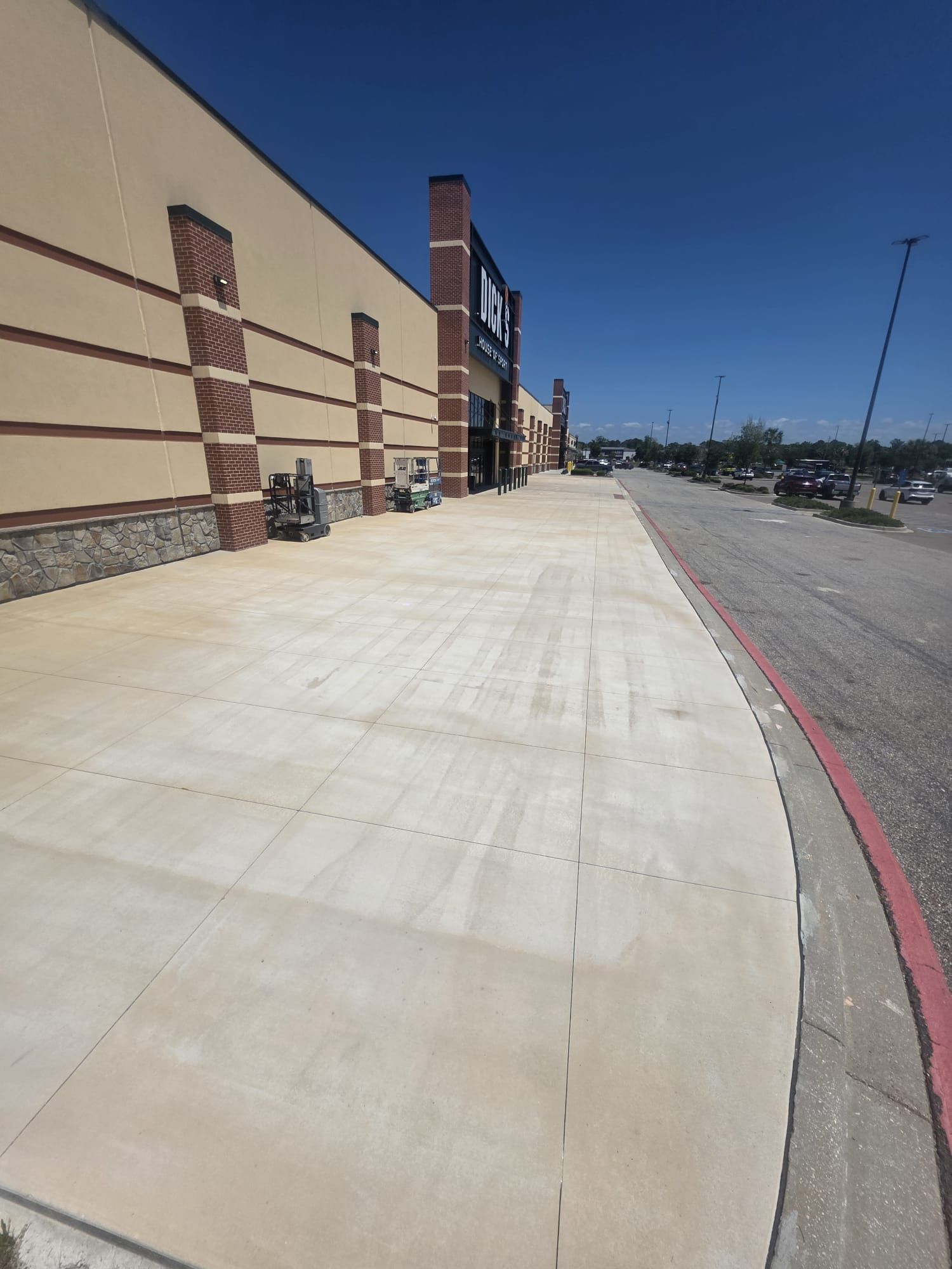 Empty sidewalk beside a storefront under a clear blue sky, with a red curb and parking lot ahead