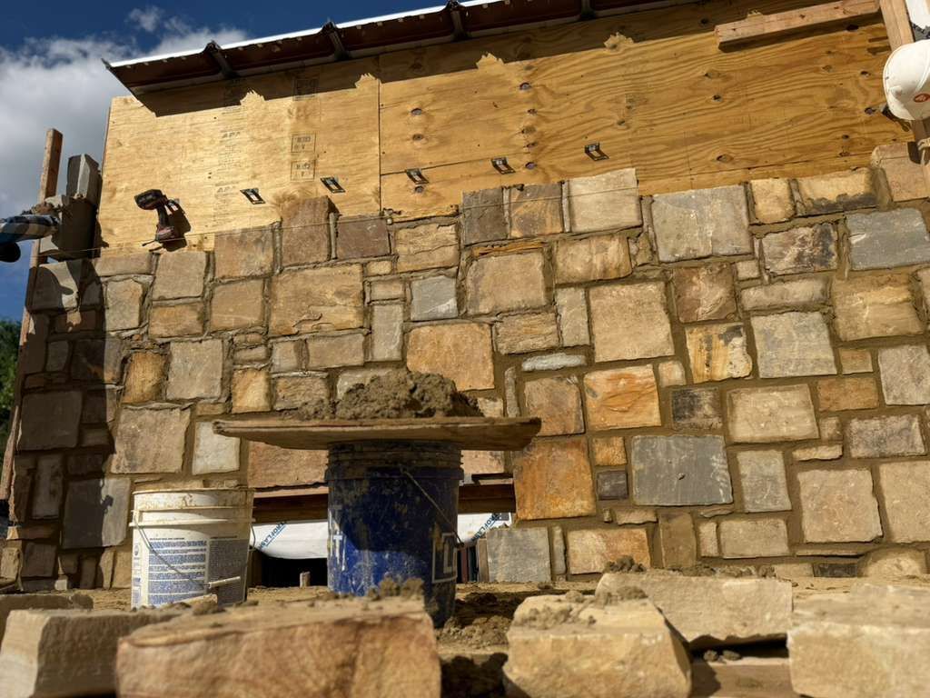 Stone wall of a rustic building with a small wooden roof and a blue barrel in front