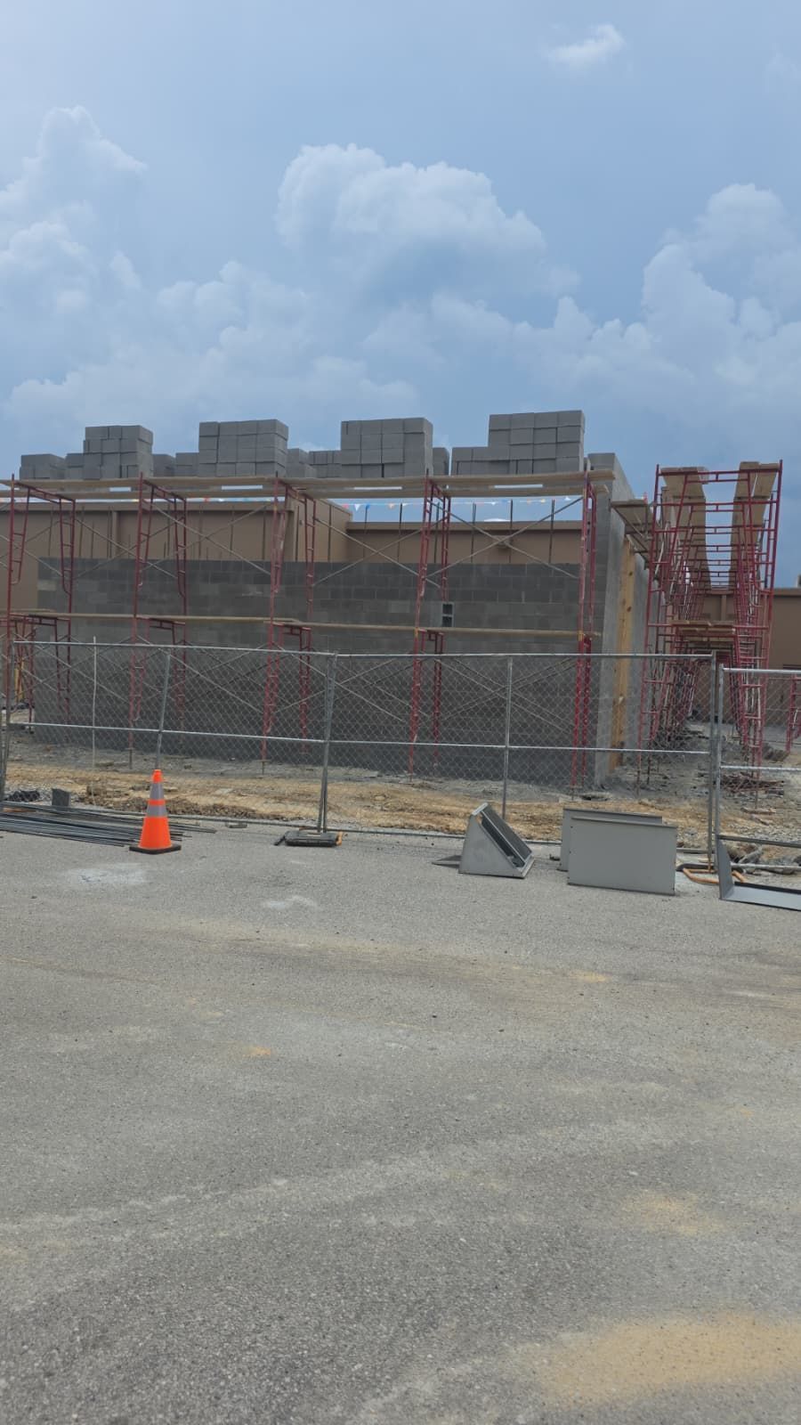 Construction site with scaffolding and concrete wall under a cloudy sky, behind a chain-link fence.