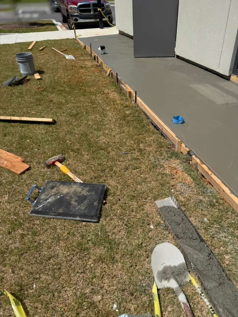 Construction tools and materials beside a building’s foundation along a grassy edge and concrete walkway