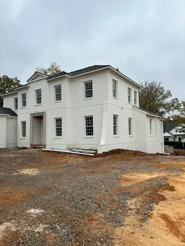 White two-story house under construction on a muddy lot, with unfinished windows and doors.