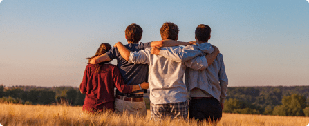 Four friends stand arm-in-arm in a sunlit field, facing a distant tree line at sunset.