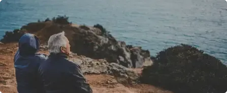 Two people in hoodies sit on a rocky cliff overlooking the ocean at sunset