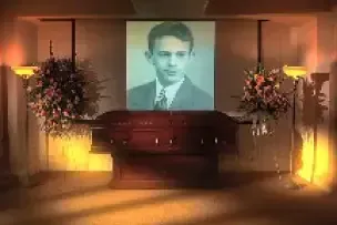 Memorial display with a portrait above a casket, flanked by floral arrangements and warm candlelight