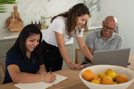 Three people collaborating around a table, one writing, one on a laptop, and the third leaning in, smiling.