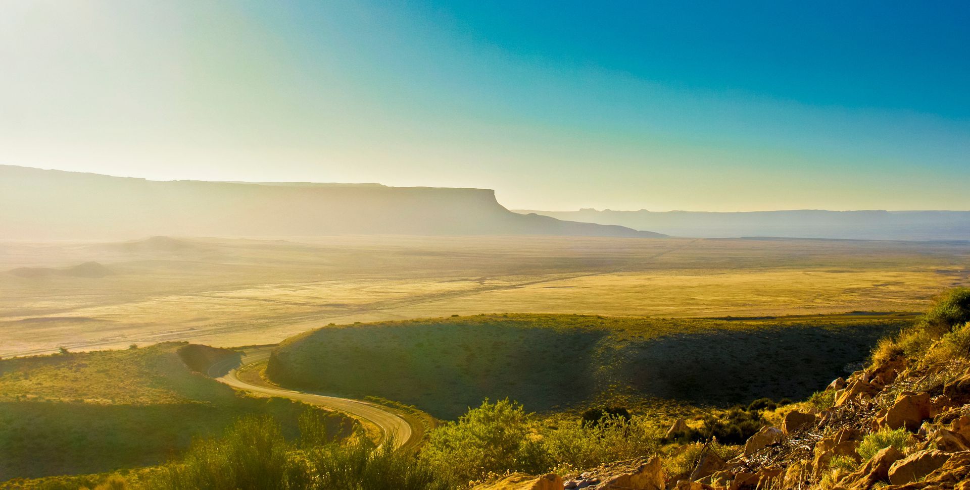 Sunlit green valley with a winding river, distant cliffs, and mist under a clear blue sky