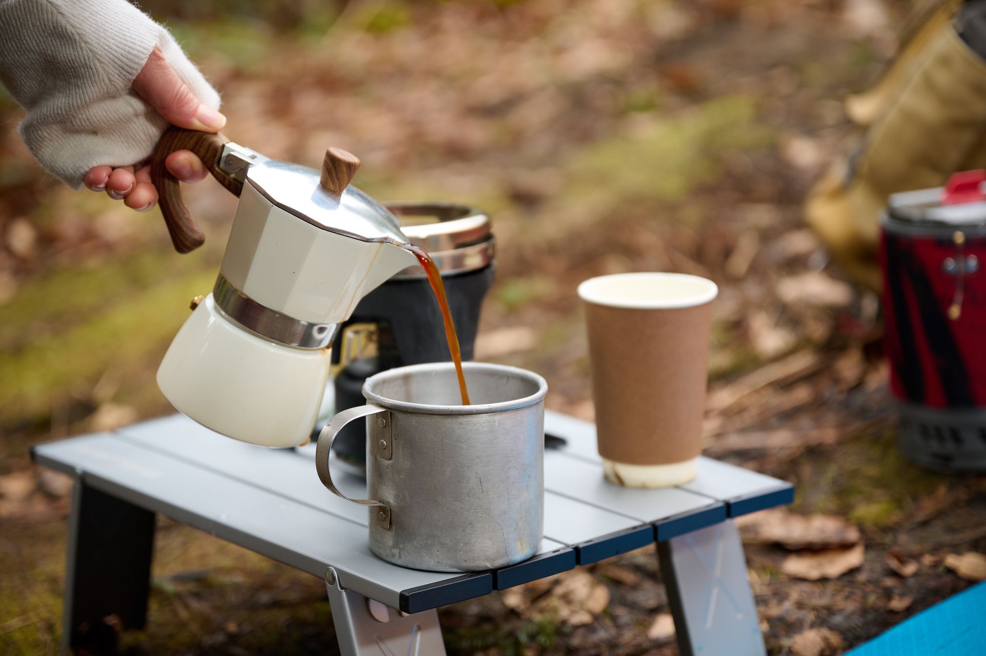Hand pouring coffee from a silver moka pot into a metal mug on a small camping table outdoors
