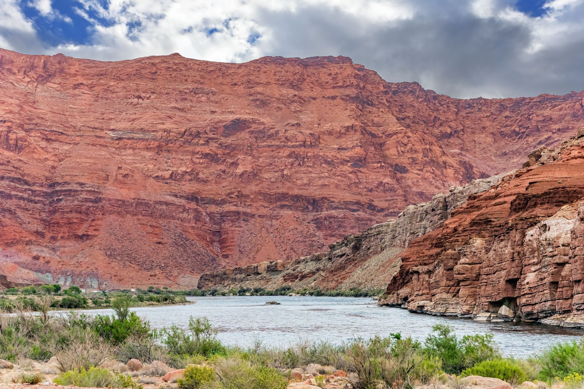Red canyon cliffs above a wide river under a cloudy sky, with sparse green shrubs in the foreground