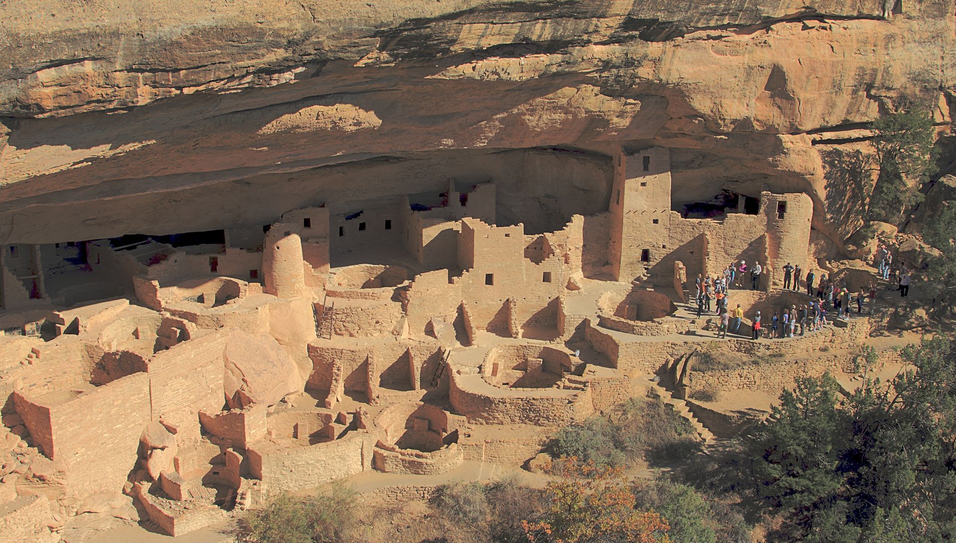 Sunlit red sandstone cliffs rise above a rocky desert landscape under a blue sky.