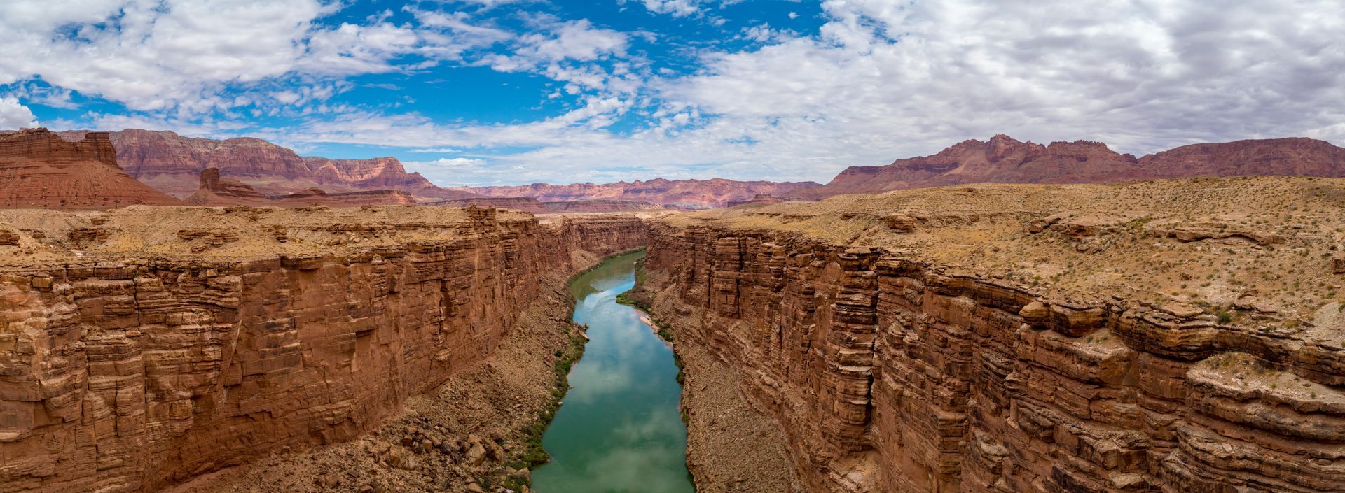 Narrow green river winding through a rocky desert canyon under a blue sky with clouds
