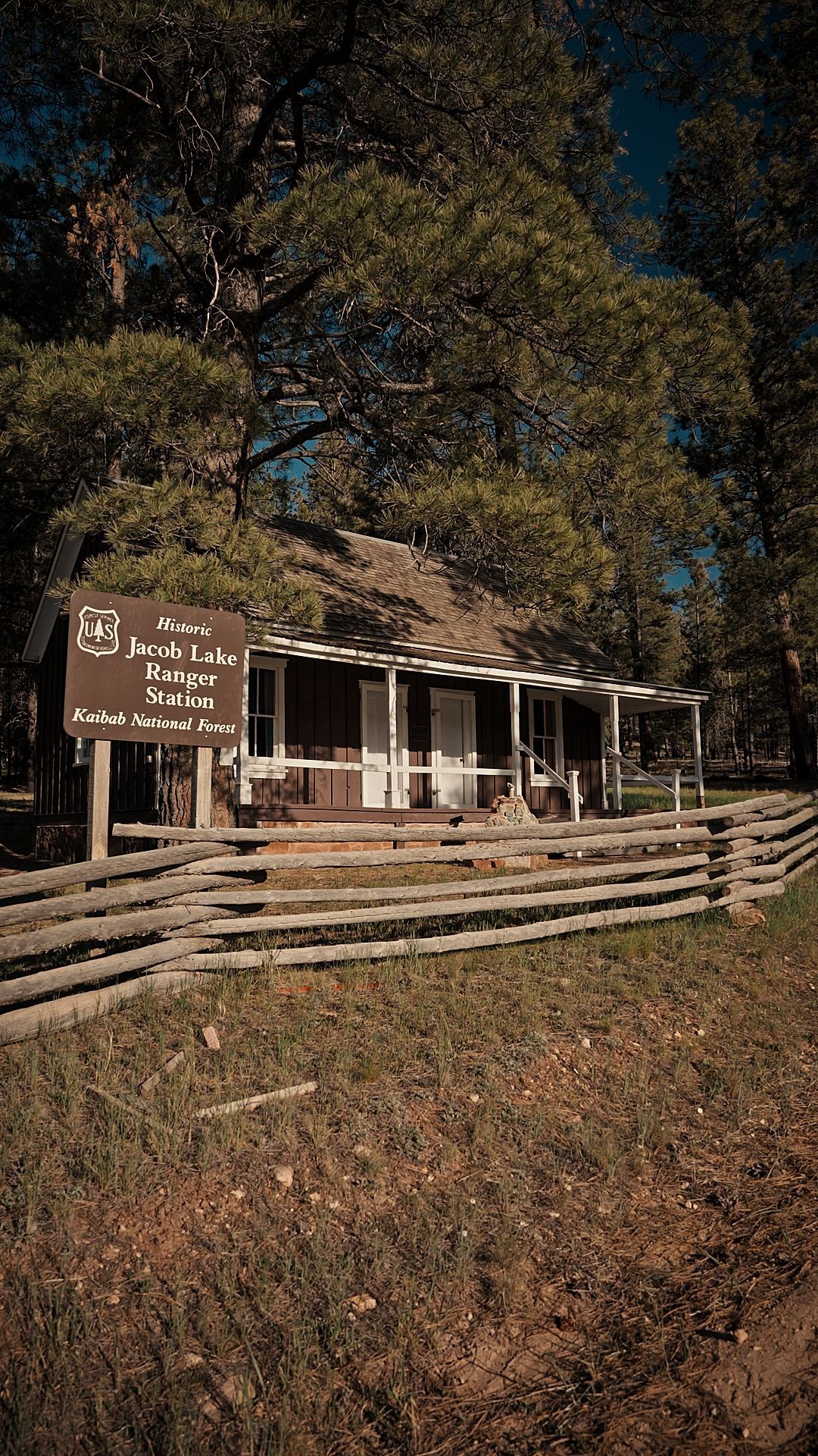 Rustic wooden cabin with porch and sign, behind a split-rail fence in a wooded area