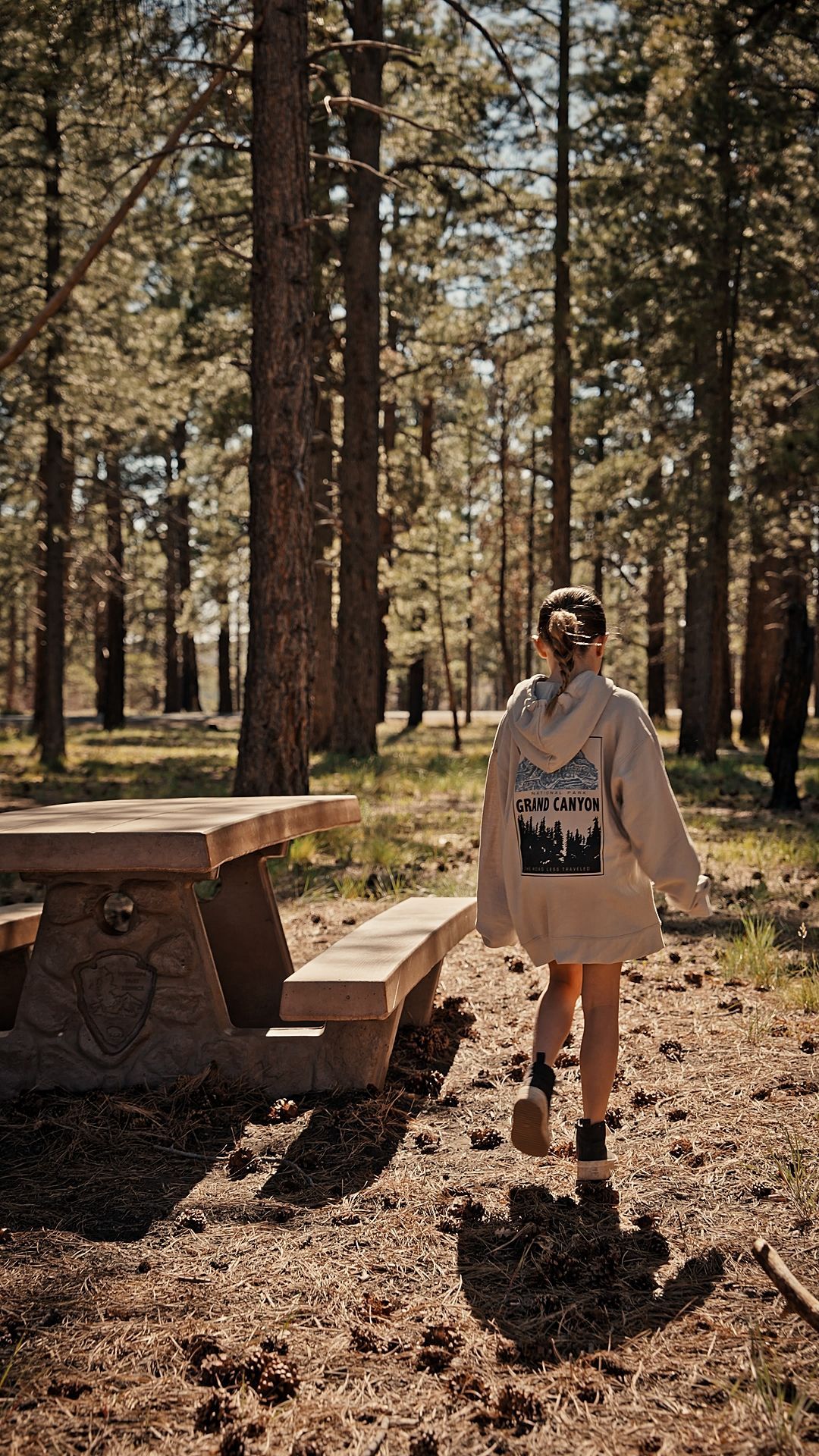 Person standing in a sunlit pine forest beside a wooden picnic table and bench