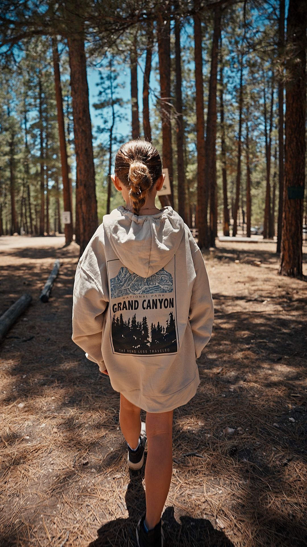 Person walking on a forest path in a white hoodie, surrounded by tall pine trees and sunlight.