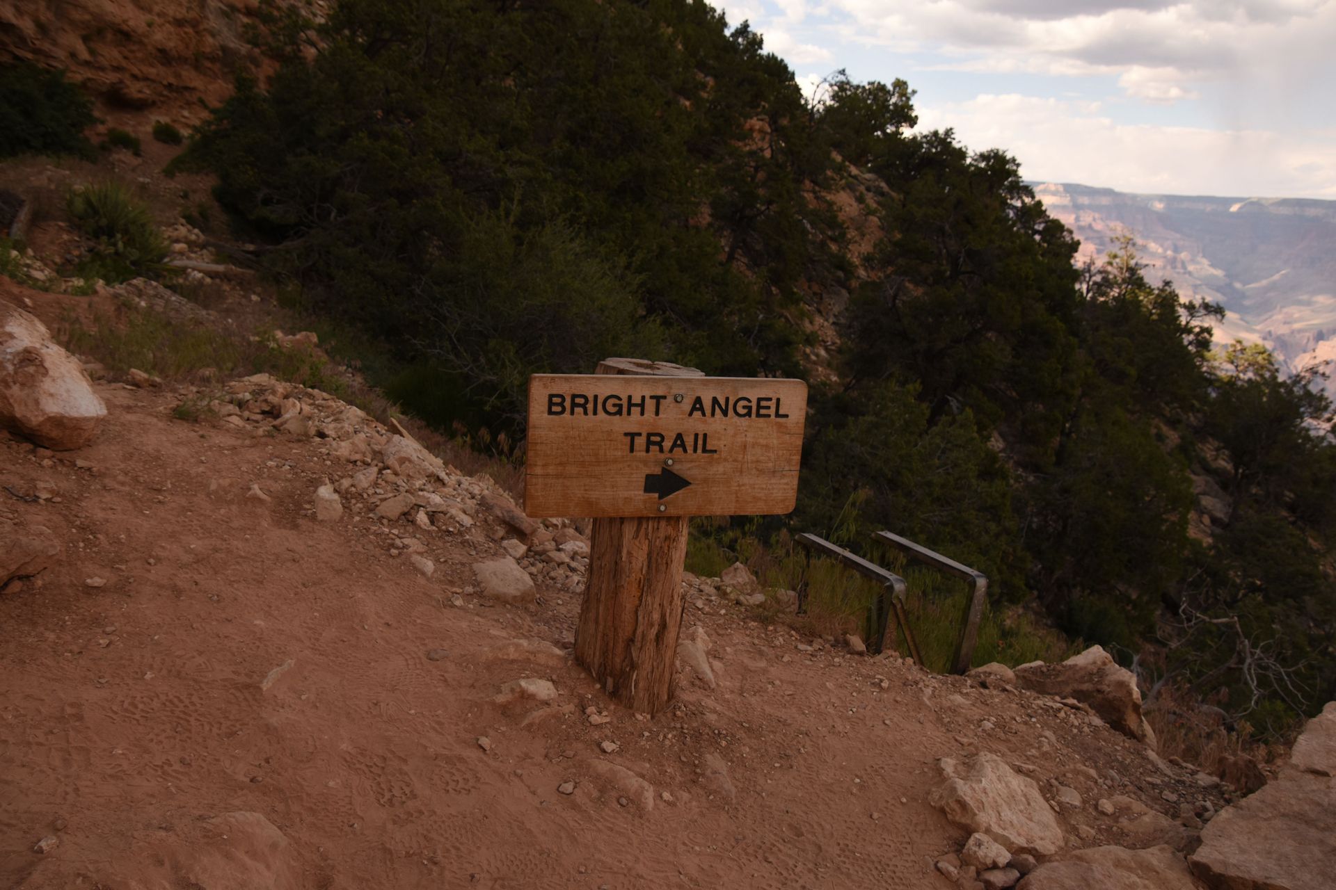 Trail sign reading “BRIGHT ANGEL TRAIL” on a rocky canyon path with trees and distant cliffs.