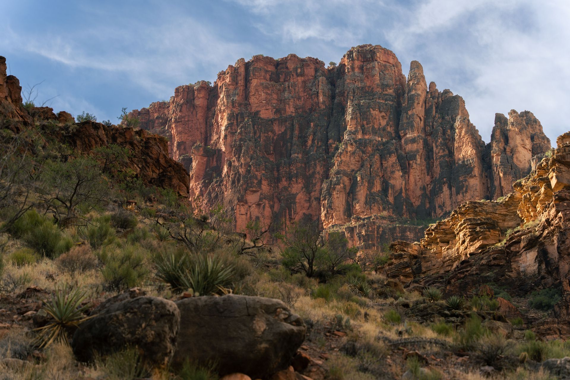 Sunlit red sandstone cliffs rise above a rocky desert landscape under a blue sky.
