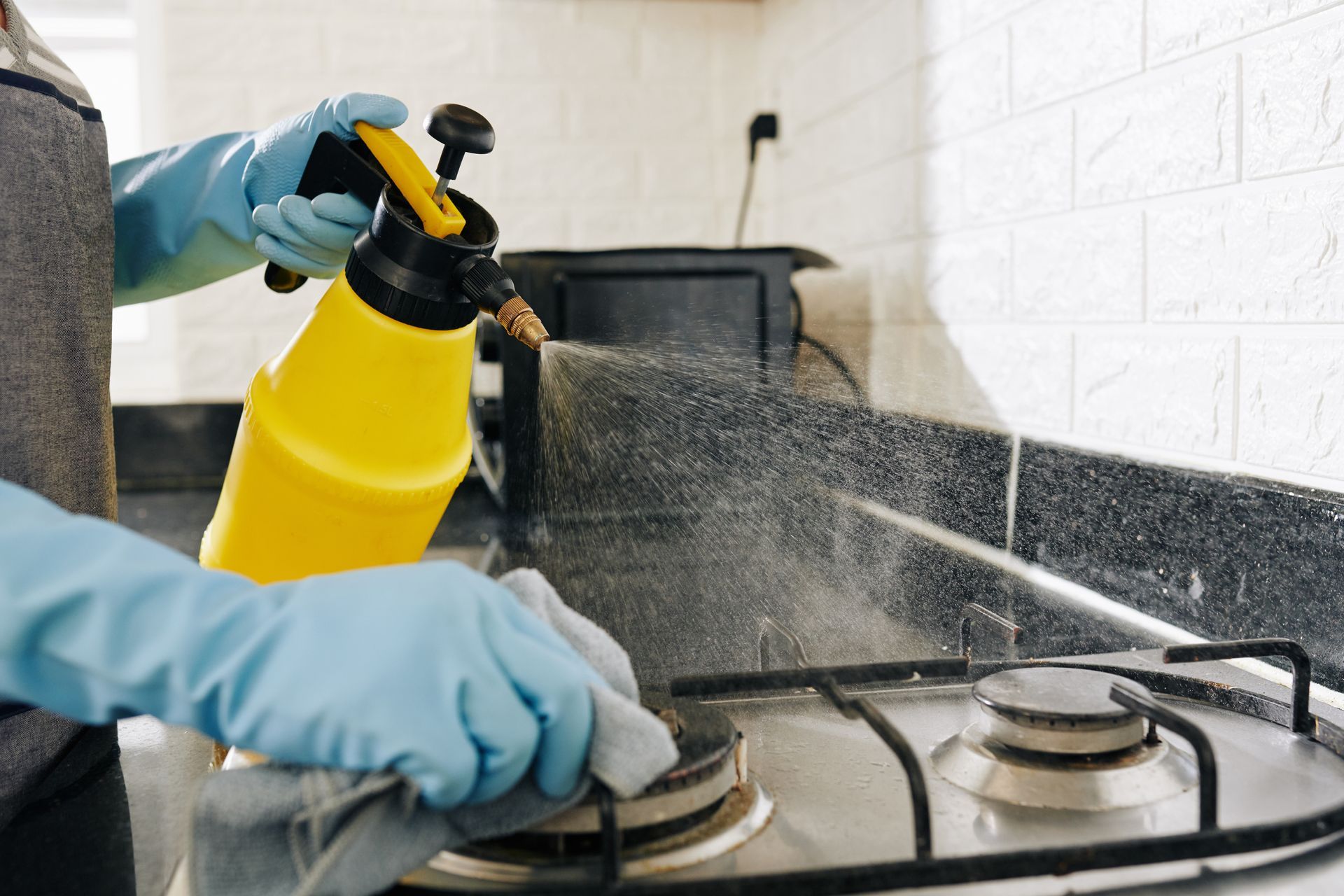 Gloved hands spray cleaner onto a gas stove in a kitchen