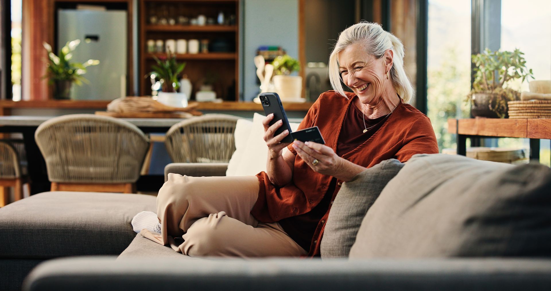 Smiling woman on a couch using a smartphone with a dog beside her in a cozy living room.
