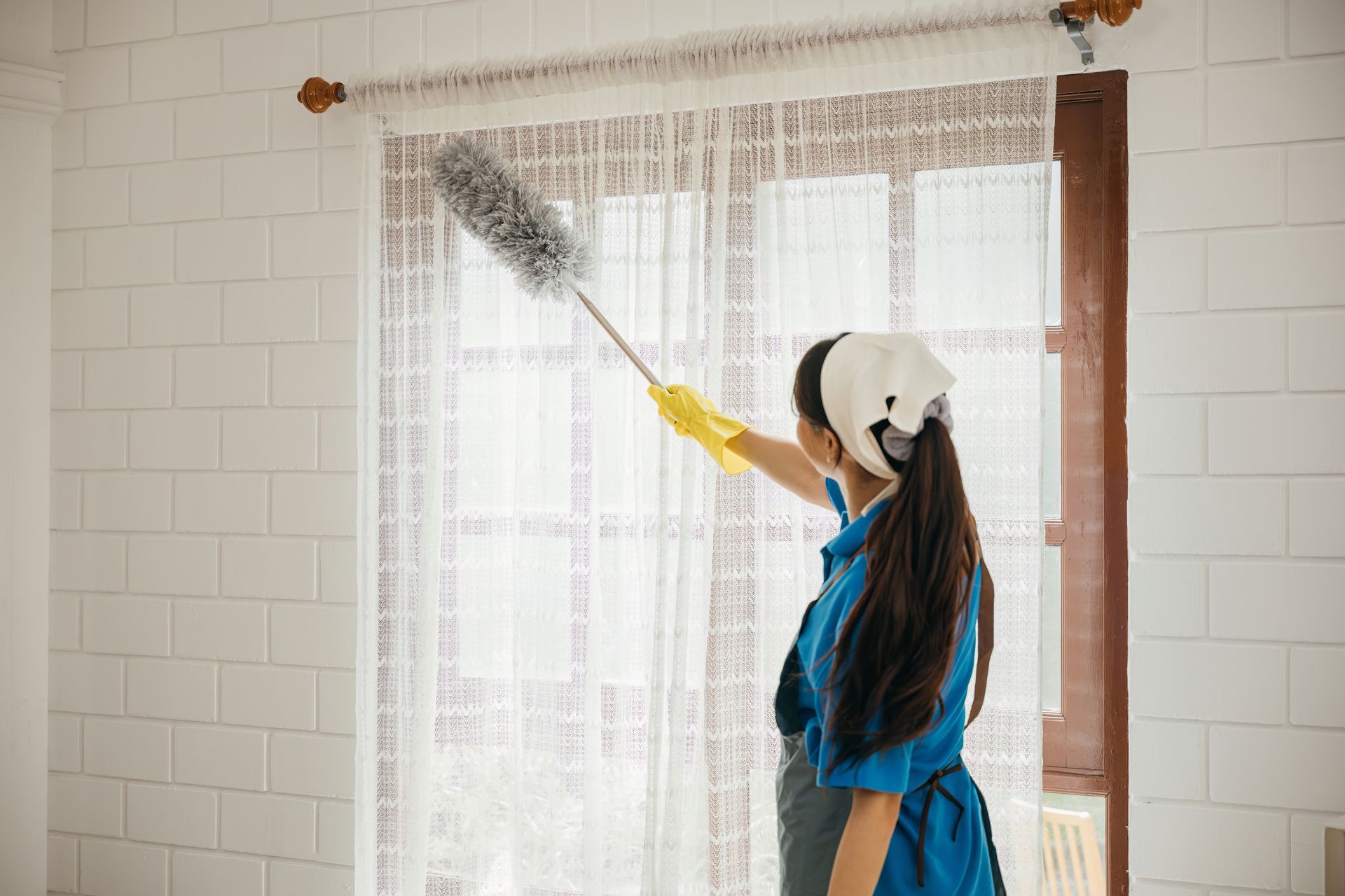 Person dusts a sheer curtain with a feather duster in a bright room