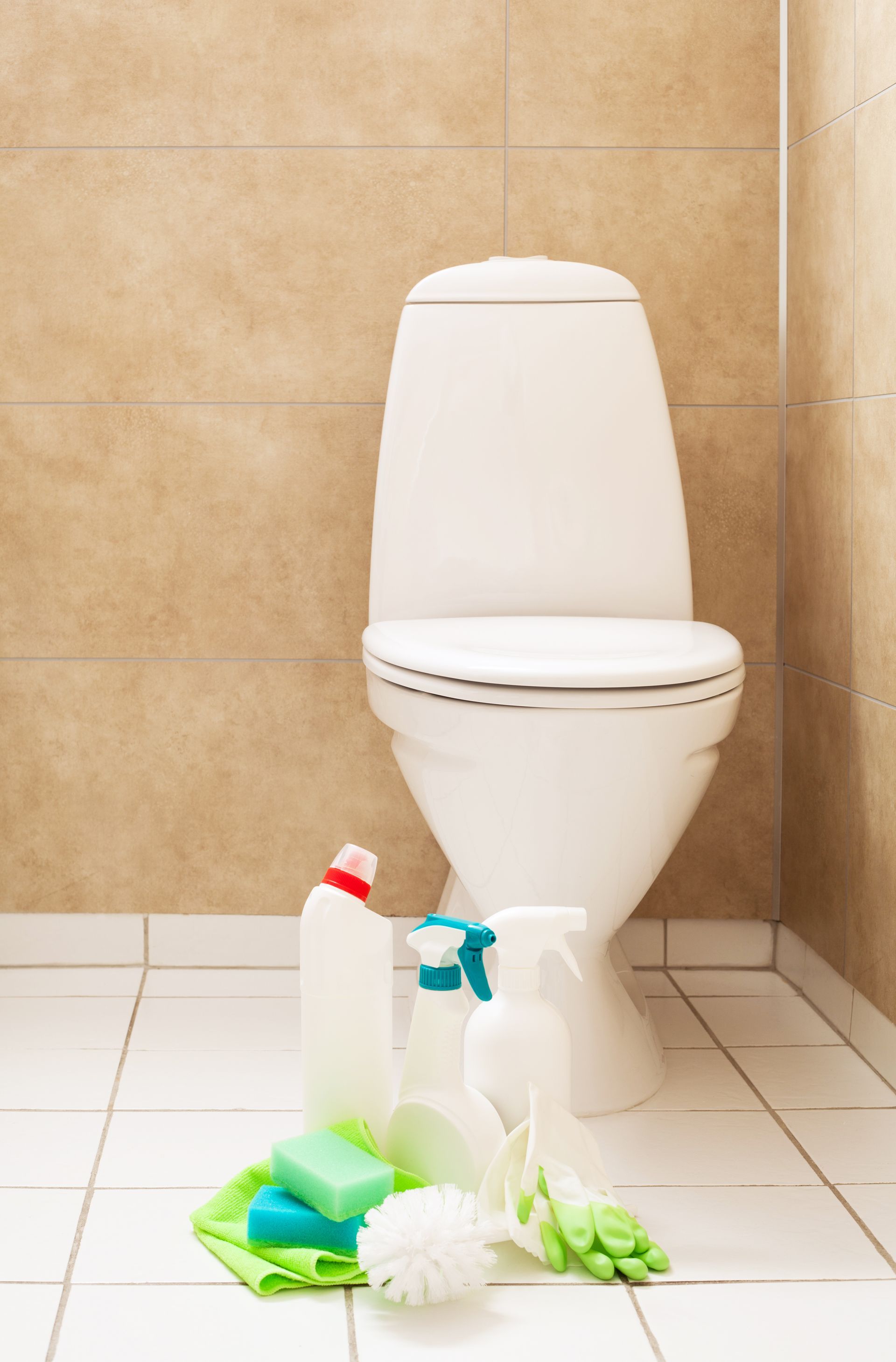 Bathroom with white toilet and cleaning supplies on the floor, including a spray bottle, brush, sponge, and gloves